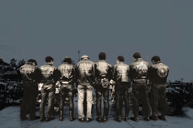 Eight riders seen from behind in matching leather jackets with back patches, grey sky