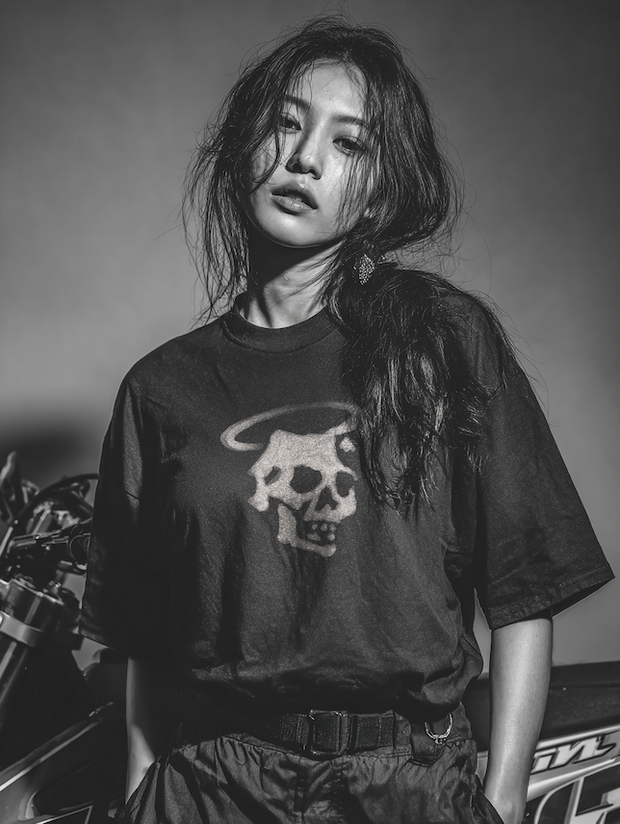 Woman with long dark hair in a black tee with a haloed skull print, motorcycle behind