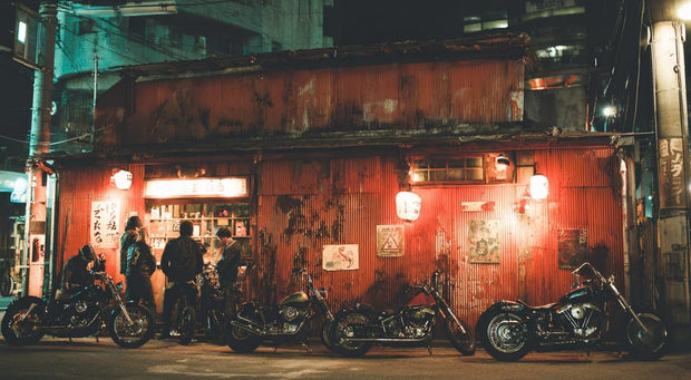 Riders standing outside a red-lit corrugated bar at night, motorcycles parked out front